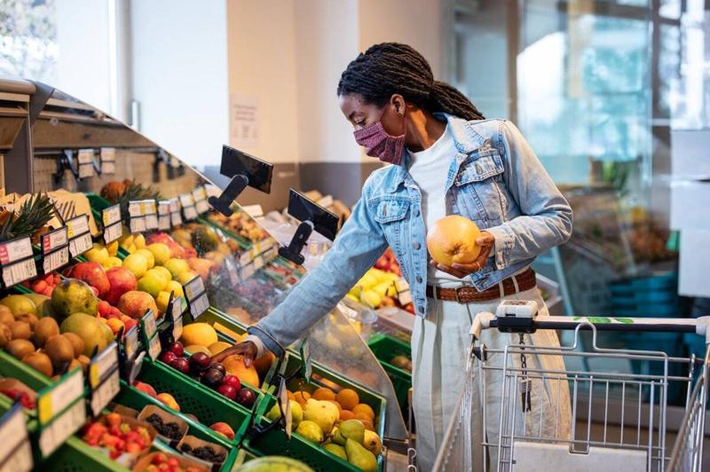 Female shopping grocery in organic shop. African woman with face mask buying fresh fruits from produce aisle in supermarket.